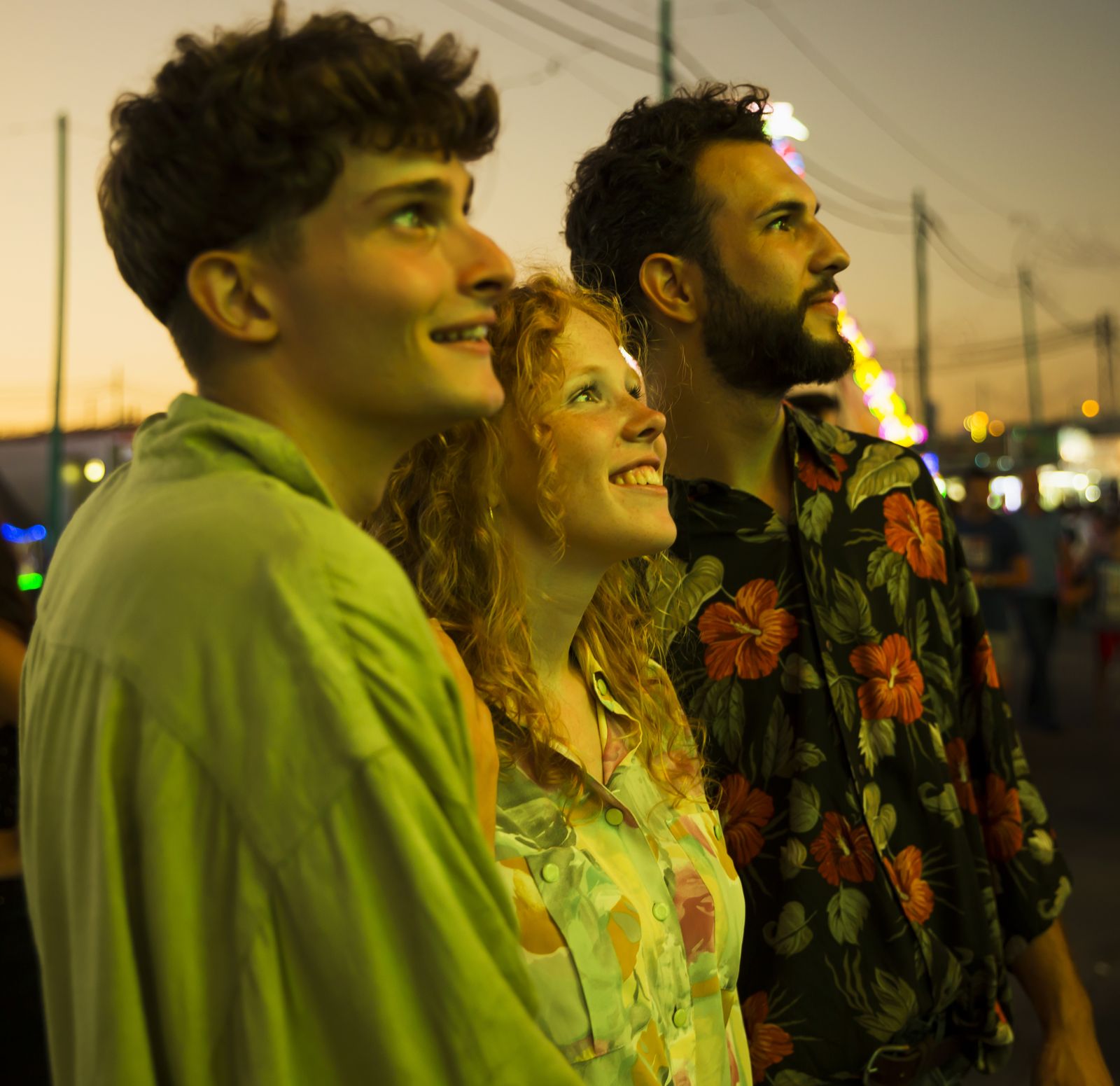Três amigos sorrindo e olhando para cima em uma feira ao entardecer