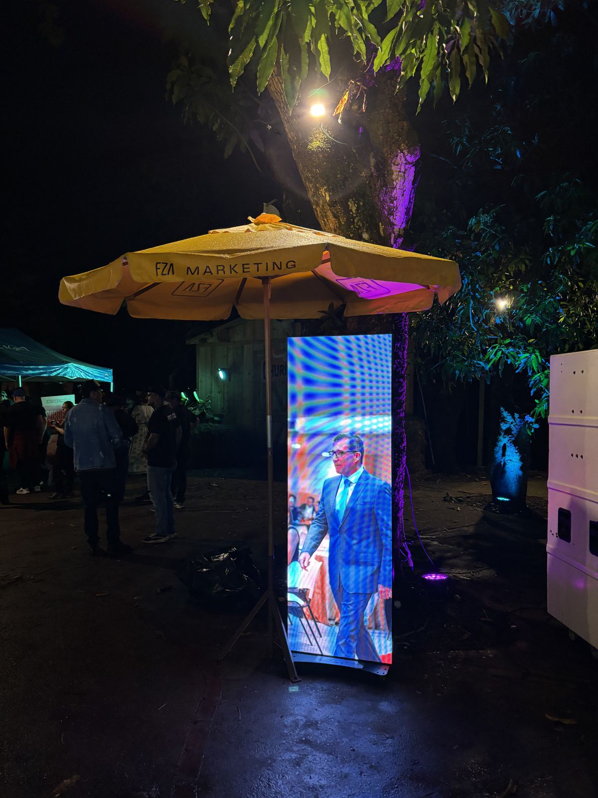 Totem de LED da Somos LED em um evento noturno, exibindo vídeo ao lado de um guarda-sol da FZA Marketing